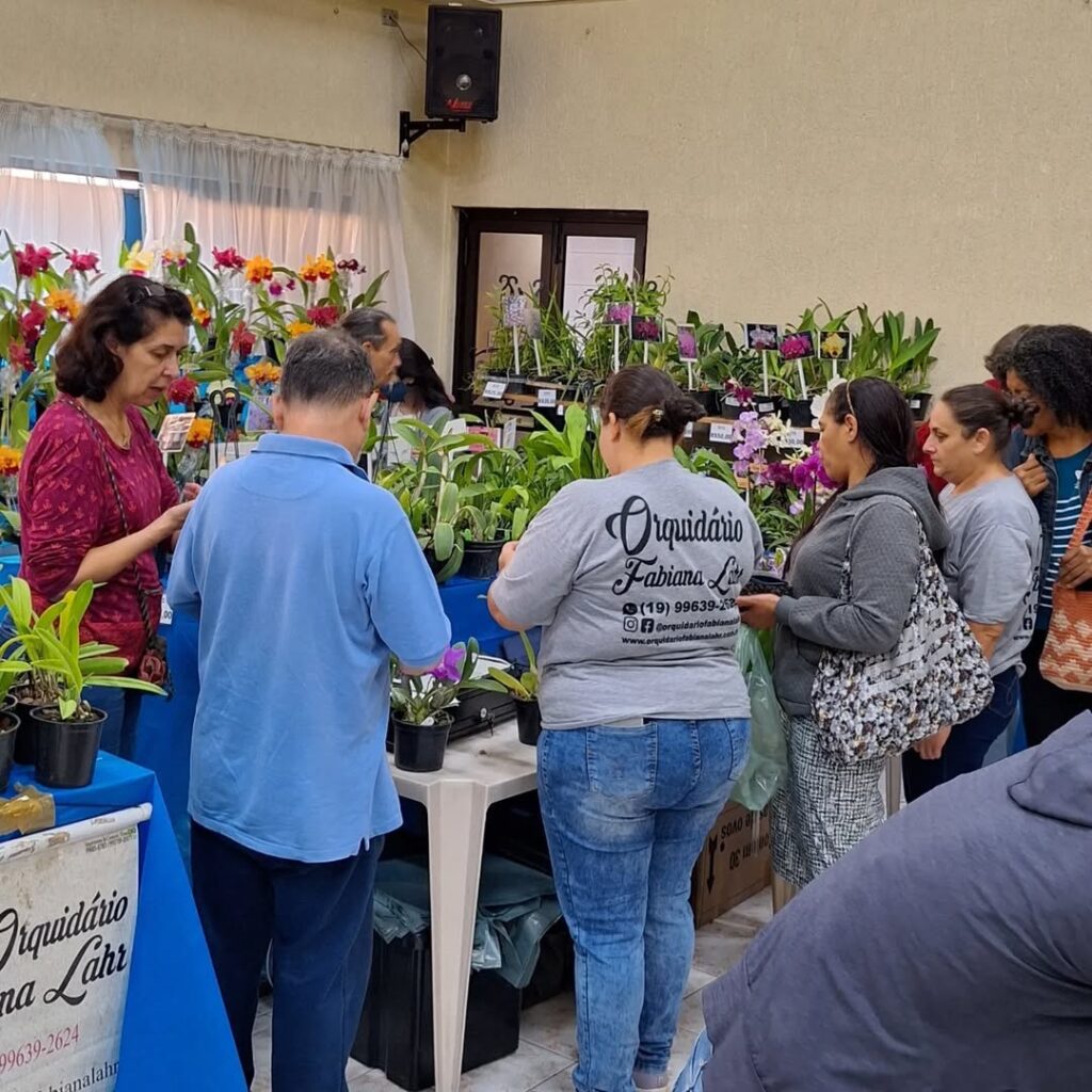 A imagem mostra uma feira ou exposição de orquídeas. Há várias pessoas olhando e comprando plantas. Há muitas orquídeas em vasos expostos em mesas. Uma mulher está usando uma camisa com o logotipo de um orquidário chamado "Orquidário Fabiana Lahr", com informações de contato. Há também uma faixa com o mesmo nome e informações de contato. O ambiente parece ser um salão ou área coberta. Há também outras plantas além de orquídeas.
