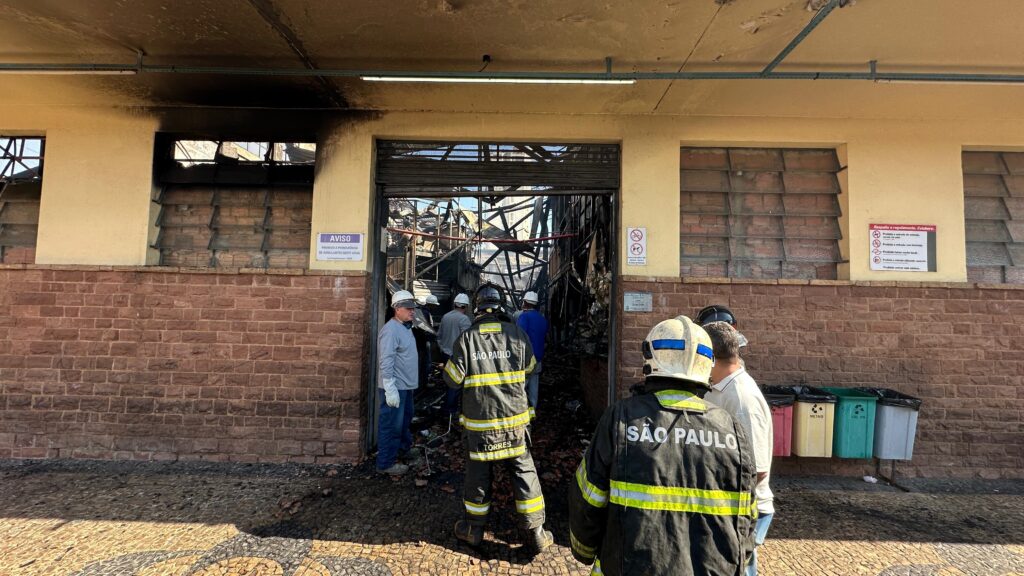 A imagem mostra uma entrada de um edifício com sinais visíveis de incêndio. O teto acima da entrada está escurecido pela fumaça e há destroços visíveis no interior. Vários bombeiros, vestidos com seus uniformes de proteção, estão em frente à entrada, alguns olhando para dentro e outros conversando. Há também alguns civis presentes. Nas paredes do edifício, há placas de aviso e sinalização. À direita da entrada, há uma fileira de lixeiras coloridas. O chão em frente ao edifício é pavimentado com pedras em um padrão circular.