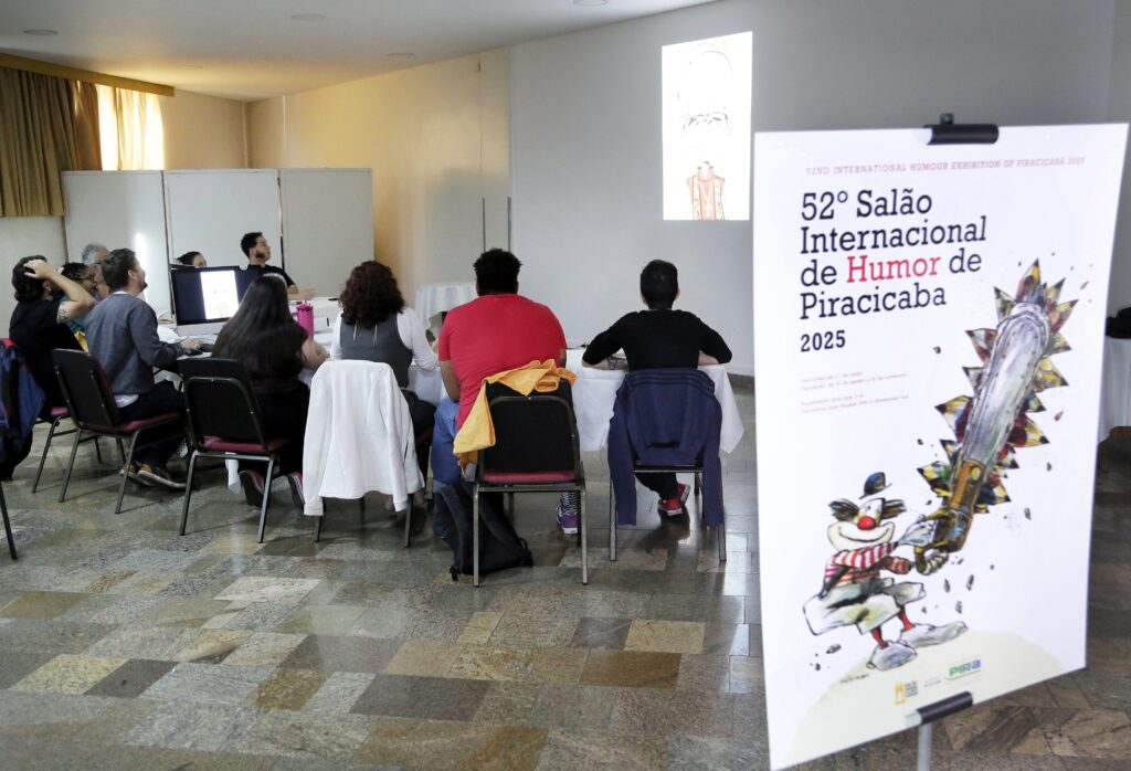 A cena se passa em uma sala de eventos com um piso de ladrilhos quadriculados em tons de bege e cinza. No centro da sala, um grupo de pessoas está reunido em torno de uma mesa coberta com toalhas brancas, participando de uma apresentação. Algumas pessoas estão sentadas, voltadas para um projetor que exibe uma caricatura no canto superior direito da sala. Há também um computador sobre a mesa, indicando que a apresentação é interativa ou baseada em mídia digital. Em primeiro plano, à direita, um grande cartaz anuncia a "52º Salão Internacional de Humor de Piracicaba 2025". O cartaz apresenta uma ilustração vibrante com uma serra elétrica estilizada e um personagem de palhaço sorridente. O texto no cartaz indica que o evento é uma exposição internacional de humor. As cadeiras ao redor da mesa são pretas com assentos e encostos acolchoados em vermelho, e algumas delas estão cobertas com casacos. O ambiente parece formal, mas com um toque de informalidade devido à natureza do evento e à disposição das pessoas