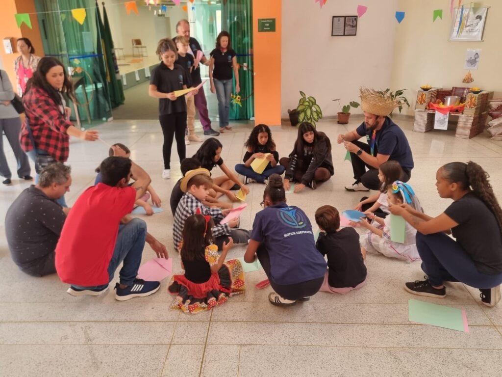 A imagem mostra um grupo de crianças e adultos reunidos em uma sala, aparentemente participando de uma atividade de artesanato. As crianças estão sentadas em círculo no chão, cada uma com uma folha de papel colorida, provavelmente fazendo dobraduras ou criando algo com o papel. Há adultos circulando as crianças, alguns parecem estar ajudando ou supervisionando a atividade. O ambiente parece ser uma escola ou centro comunitário, com decoração festiva, incluindo bandeirinhas e um chapéu de palha típico de festas juninas. Os adultos e as crianças parecem estar em um ambiente descontraído e amigável, interagindo entre si. A atmosfera geral sugere uma atividade lúdica e educativa.