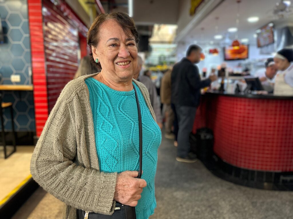 A imagem mostra uma mulher mais velha sorrindo para a câmera em um ambiente interno, possivelmente um mercado ou praça de alimentação. Ela veste um suéter azul com um padrão de cabos intricado e um cardigã bege sobre ele. Um pequeno bolsa preta com zíper está pendurada em seu ombro. Seu cabelo é curto e marrom, e ela usa um brinco verde em sua orelha esquerda. O fundo está desfocado, mas mostra outras pessoas em uma fila em frente a um balcão vermelho com azulejos. Há também uma parede com um padrão de hexágono azul e vermelho à esquerda da mulher. A iluminação é brilhante, sugerindo um ambiente bem iluminado. A mulher parece relaxada e feliz.