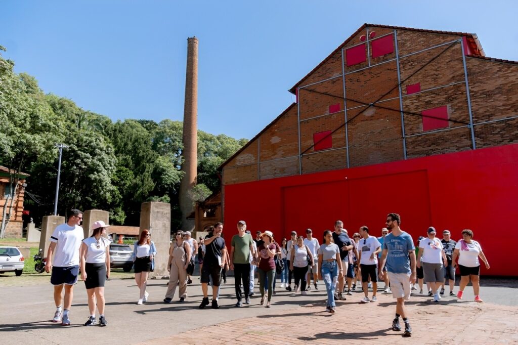 A imagem mostra um grupo de pessoas caminhando em direção à câmera em um dia ensolarado. Estão em frente a um prédio de tijolos com uma grande parede vermelha e um telhado inclinado. Há uma chaminé alta e estreita de tijolos ao fundo, e árvores verdes cercam a área. As pessoas estão vestidas com roupas casuais de verão, e algumas parecem estar em um passeio guiado. O chão é pavimentado com tijolos.