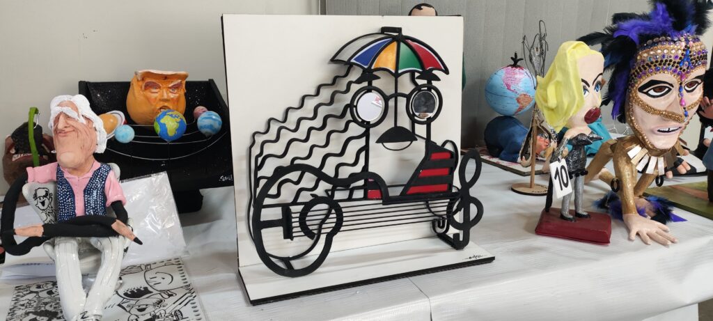 A imagem mostra uma variedade de esculturas e artes em uma mesa. Há uma escultura de papel machê de um homem idoso sentado em uma cadeira, uma representação de um sistema solar com um busto de Donald Trump como o sol, uma escultura de metal que parece um músico com um guarda-chuva, e várias outras figuras menores, incluindo uma que parece ser uma representação de Madonna. As esculturas são vibrantes e coloridas, com estilos e materiais variados. A mesa parece estar em um ambiente de exibição ou concurso, sugerido pela presença de um número "10" em uma placa próxima a uma das esculturas. O fundo é uma parede simples e clara.