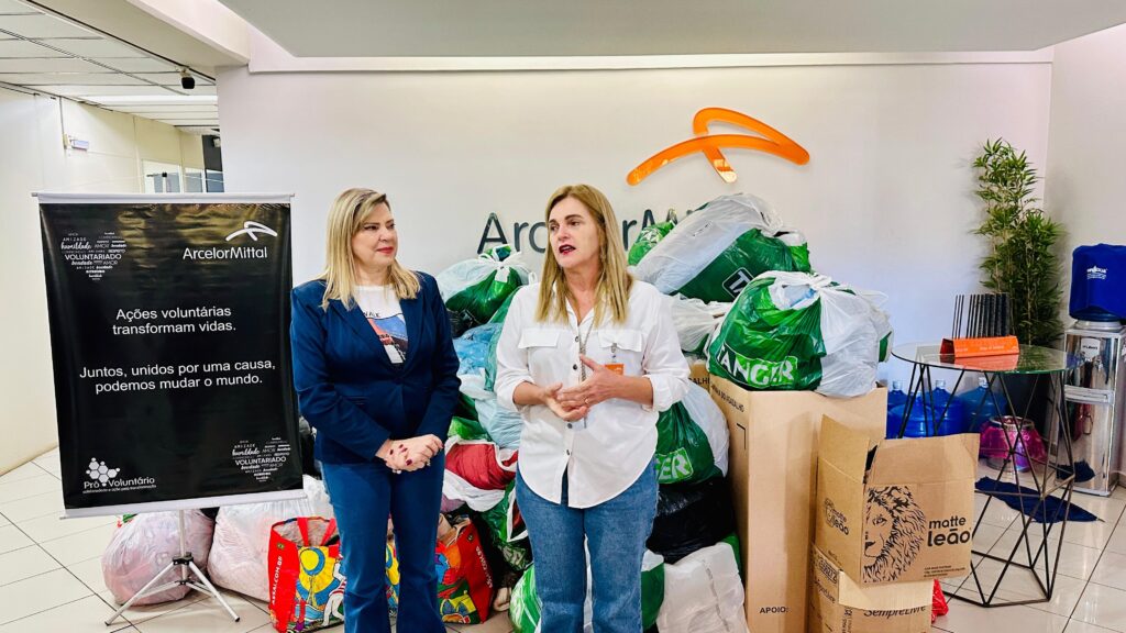 A imagem mostra duas mulheres em pé na frente de uma pilha de sacos de compras e caixas cheias de roupas doadas. Elas estão em um ambiente de escritório, com uma placa da ArcelorMittal ao fundo. A placa exibe o slogan "Ações voluntárias transformam vidas. Juntos, unidos por uma causa, podemos mudar o mundo." As mulheres parecem estar envolvidas em uma iniciativa de caridade ou trabalho voluntário. Uma delas está falando, enquanto a outra ouve atentamente. A atmosfera é formal, mas acolhedora. A imagem sugere uma ação de responsabilidade social da empresa ArcelorMittal, onde funcionários doaram roupas para uma causa beneficente.