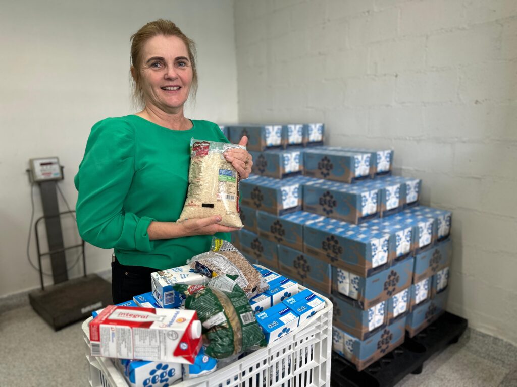 A imagem mostra uma mulher de meia-idade, com cabelo castanho claro e olhos claros, sorrindo para a câmera. Ela está usando uma blusa verde e está segurando um pacote de arroz. Ao seu lado, há um carrinho de plástico cheio de vários itens de mantimentos, incluindo leite e feijão. Ao fundo, há pilhas de caixas de leite empilhadas contra uma parede de tijolos brancos. A cena parece ser um depósito ou armazém. A mulher parece estar envolvida em alguma atividade de doação ou distribuição de alimentos. A iluminação é suave e natural, e a imagem geral é clara e bem focada.