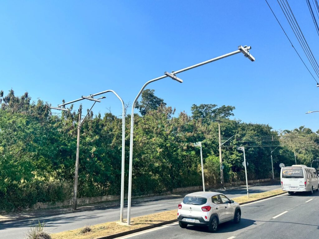 A imagem mostra uma rua com duas faixas de tráfego, ladeada por uma área gramada e uma densa vegetação. O céu está limpo e azul. No lado esquerdo da estrada, há dois postes altos com braços metálicos que se estendem sobre a rua, equipados com o que parecem ser câmeras ou sensores. Um dos postes é mais fino e curvado, enquanto o outro é mais robusto e reto. Na faixa da direita, um carro branco compacto está em movimento. Mais adiante, um ônibus branco com a palavra "Campestre" escrita na traseira também segue pela mesma via. A estrada é bem conservada, com marcações de faixa brancas visíveis. O ambiente geral sugere uma área urbana ou suburbana com bastante verde.