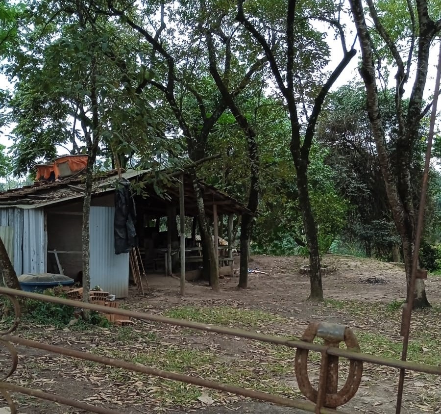 A imagem mostra uma pequena estrutura de madeira e metal, que parece ser um galpão ou abrigo, situado em um terreno arborizado. A estrutura é simples, com um telhado de madeira e paredes de metal ondulado. Há alguns objetos visíveis dentro e ao redor da estrutura, incluindo uma lona preta pendurada em uma das paredes e alguns tijolos no chão. O terreno é de terra batida e coberto por folhas secas. Árvores de diferentes tamanhos e tipos cercam a estrutura, criando um ambiente rural e sombreado. Um pedaço de cerca de metal enferrujado com um ferradura como detalhe está em primeiro plano, parcialmente obstruindo a vista. O cenário geral sugere uma área rural ou uma propriedade em uma zona rural.