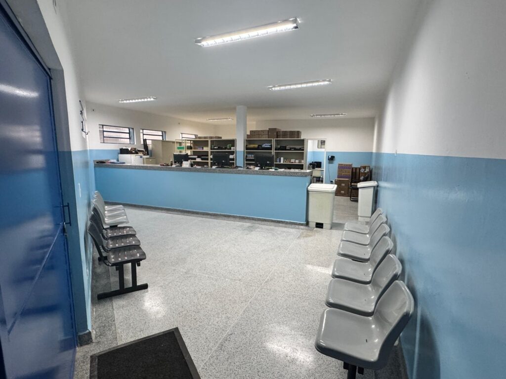 A imagem mostra um corredor de espera em um local que parece ser uma clínica ou farmácia. * Há uma porta azul grande à esquerda e uma parede azul clara com uma linha branca no meio. * No lado direito, há uma fila de cadeiras de espera cinzas. * Ao fundo, há um balcão azul claro com computadores e prateleiras atrás dele, onde estão armazenados produtos. * O chão é de granilite claro. * HáHá também algumas lixeiras e caixas empilhadas em um canto.