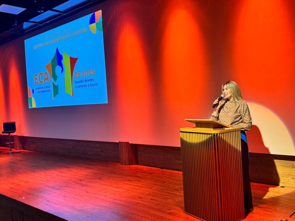 A imagem mostra uma mulher loira falando em um microfone, de pé atrás de um púlpito de madeira em um palco. Ela está usando uma blusa estampada e calças azuis. Atrás dela, em uma tela grande, está escrito em português: "I Seminário Municipal do SISTEMA DE GARANTIA DE DIREITOS ECA Estatuto da Criança e do Adolescente 35 anos Garantir direitos é construir o futuro". O palco e o fundo estão iluminados com luzes vermelhas e laranjas vibrantes, criando uma atmosfera de evento. À esquerda, há uma cadeira de palco e à direita, um pequeno tronco de madeira.
