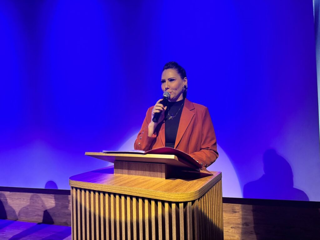 A imagem mostra uma mulher de cabelo escuro preso num coque, vestindo um blazer laranja e blusa preta, falando ao microfone atrás de um púlpito de madeira. O fundo é uma parede azul vibrante. Ela parece estar dando um discurso ou apresentação. A iluminação é suave e focada principalmente na mulher, criando um contraste com o fundo. A expressão facial dela sugere seriedade e concentração. O ambiente parece formal, talvez um auditório ou palco.