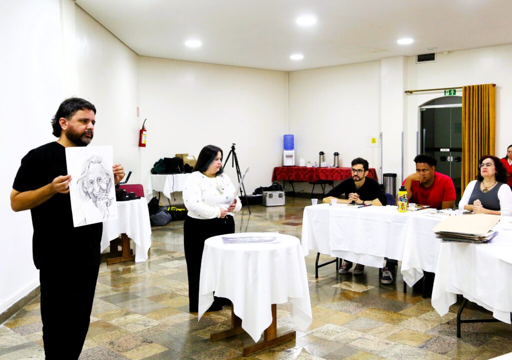 A imagem mostra um grupo de pessoas em uma sala, possivelmente para uma aula ou apresentação. No lado esquerdo, um homem de camiseta preta está em pé, segurando um desenho de um rosto envelhecido, feito a lápis. Ele parece estar apresentando o desenho para o grupo. À sua direita, uma mulher de camiseta branca com detalhes coloridos está em pé, olhando para o desenho. Atrás dela, há um tripé com uma câmera ou projetor. Ao fundo, há mesas cobertas com toalhas brancas, e algumas pessoas estão sentadas a elas, participando do evento. Uma mesa ao fundo está coberta com uma toalha vermelha e tem uma garrafa de água e outros itens. A sala tem paredes brancas e um piso com um padrão de ladrilhos coloridos. Há também extintores de incêndio nas paredes e luminárias no teto. A iluminação é clara, sugerindo que o evento está acontecendo durante o dia ou em um local bem iluminado.