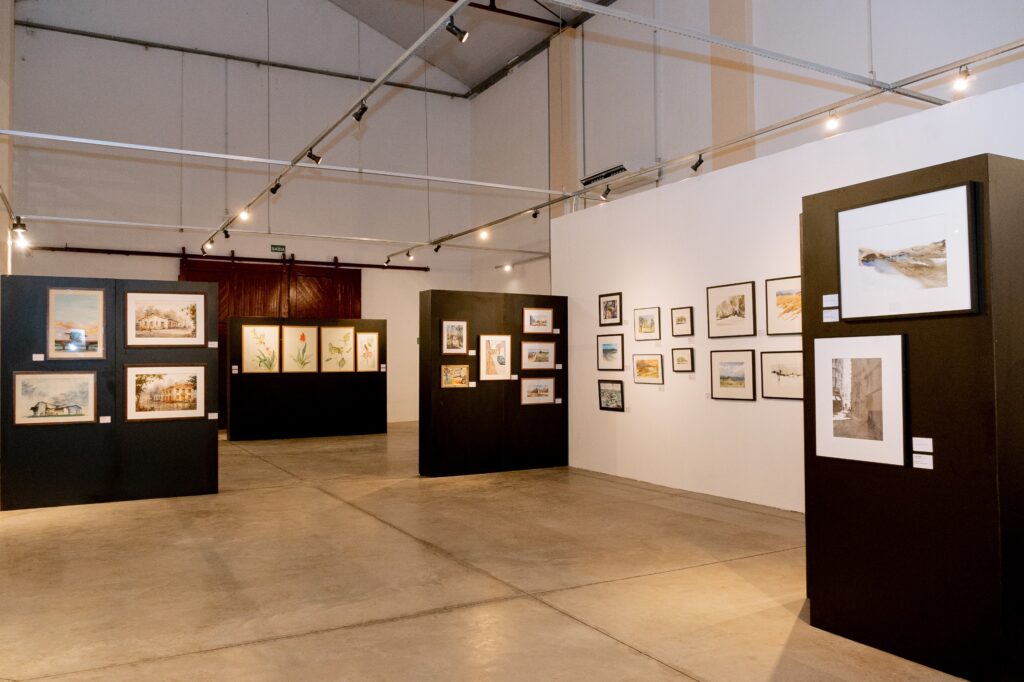 A imagem mostra uma galeria de arte com várias pinturas expostas em paredes e painéis pretos. As paredes são brancas e o chão é de concreto polido. As pinturas são principalmente aquarelas e parecem retratar paisagens e cenas urbanas. A iluminação é suave e difusa, criando uma atmosfera calma e contemplativa. A galeria parece ser um espaço amplo e arejado, com um teto alto e vigas expostas. Há uma porta de madeira escura visível ao fundo. No geral, a imagem transmite uma sensação de serenidade e elegância.