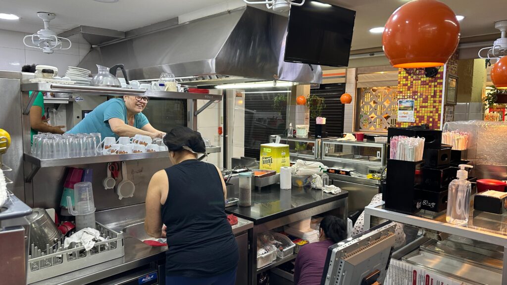 A imagem mostra o interior de um estabelecimento, possivelmente um café ou restaurante, com foco na área de serviço e preparo. Vemos duas mulheres trabalhando: uma, sorridente e usando uma blusa azul, está atrás de um balcão, manuseando canecas e copos; a outra, de costas e vestindo uma regata preta, está em frente a uma pia, aparentemente lavando algo. O ambiente é bem iluminado, com destaque para o exaustor de aço inoxidável acima da área de trabalho. Há prateleiras com pilhas de copos e canecas, além de utensílios de cozinha, como tesouras e conchas. No balcão, observamos diversos itens, incluindo uma caixa de um produto chamado "Travnic", rolos de papel, recipientes com canudos e um frasco de álcool em gel. Ao fundo, uma parede com azulejos coloridos e um calendário chamam a atenção, assim como duas luminárias esféricas laranjas suspensas. Uma terceira pessoa, usando uma blusa roxa, está agachada em segundo plano, também trabalhando.