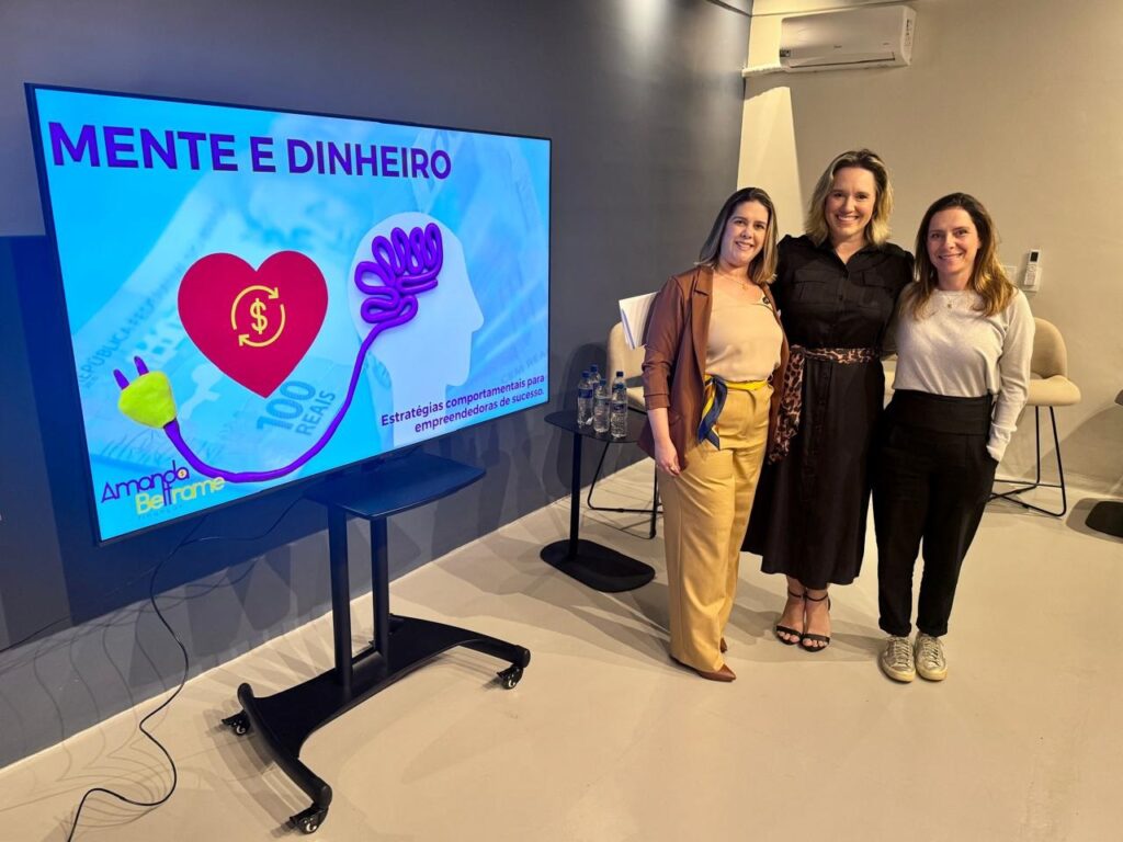 A imagem mostra três mulheres posando para uma foto em frente a uma tela grande. A tela exibe o título "MENTE E DINHEIRO" em letras roxas grandes. Abaixo do título, há um gráfico que mostra um coração vermelho com um símbolo de dólar ($) dentro, conectado por um cabo a uma silhueta de cabeça com um cérebro roxo. Há também o texto "Estratégias comportamentais para empreendedoras de sucesso" na tela, juntamente com a assinatura "Amanda Beltrame". As três mulheres estão posicionadas lado a lado. A mulher à esquerda usa uma blusa bege e uma jaqueta marrom, com calças amarelas e um lenço azul e amarelo na cintura. A mulher do meio veste um vestido preto com um cinto estampado de oncinha. A mulher à direita está de blusa cinza e calças pretas. Todas sorriem para a câmera. Ao fundo, à esquerda, há uma mesa com garrafas de água e uma cadeira. Ao fundo, à direita, há um ar condicionado na parede e cadeiras. A iluminação da sala é clara e moderna.