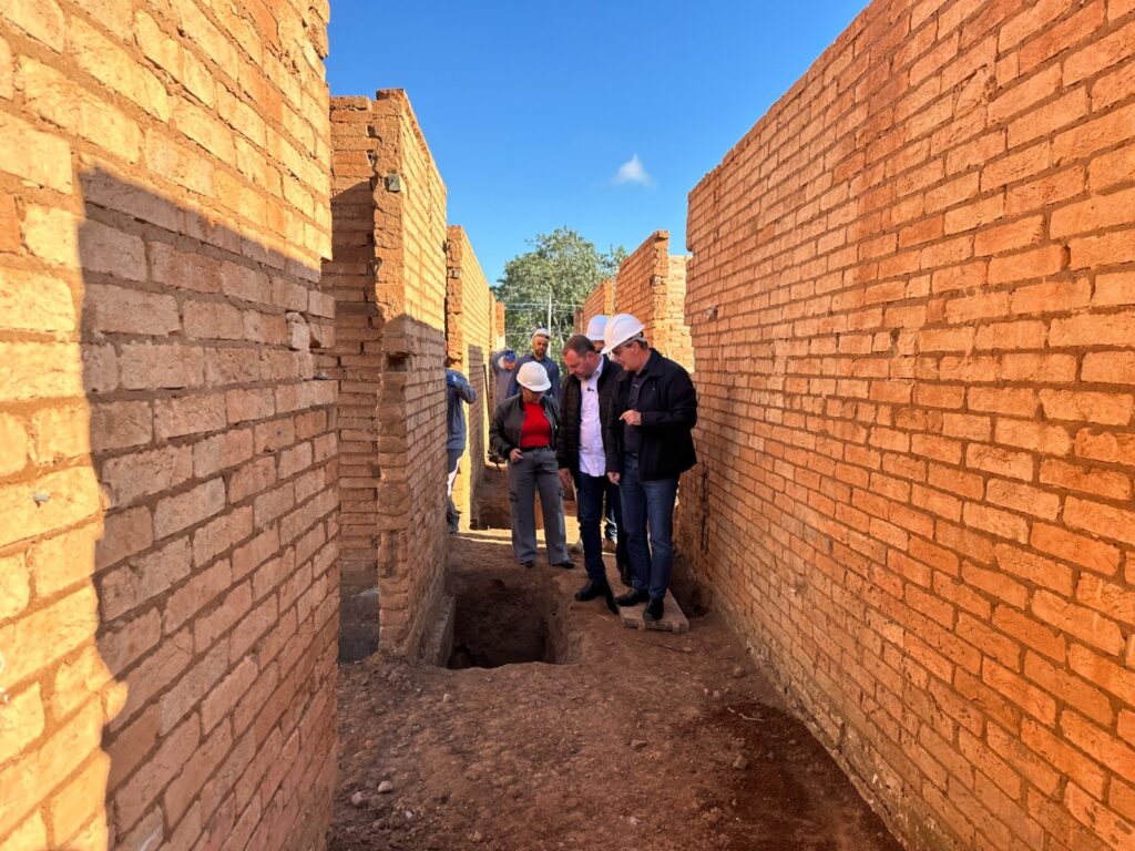 A imagem mostra um grupo de pessoas, aparentemente profissionais da construção civil, inspecionando uma obra em construção. Eles estão usando capacetes brancos e estão reunidos em um corredor estreito formado por paredes de tijolos de terracota. O corredor é parcialmente escavado, revelando solo. A iluminação sugere que a foto foi tirada durante o dia, em um local ensolarado. O céu é azul claro e sem nuvens. O ambiente parece ser rural ou em uma área com pouca urbanização. Os tijolos parecem ser feitos de barro cozido, e a construção está em fase inicial. A cena transmite uma sensação de trabalho em andamento e planejamento de construção.