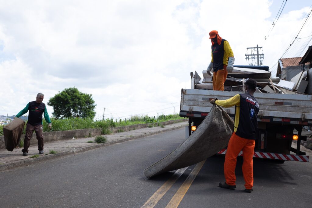 A imagem mostra três trabalhadores de limpeza pública em uniforme, coletando lixo em uma rua. Um trabalhador está carregando um objeto grande e pesado, que parece ser um colchão velho, enquanto outro coleta um saco de lixo. Um terceiro trabalhador está carregando um pedaço de entulho. Todos eles estão vestindo uniformes de segurança vibrantes, incluindo jaquetas e calças laranja e amarelas. O uniforme inclui também capacetes e luvas. O lixo está sendo carregado em um caminhão de lixo branco. O caminhão está parcialmente carregado com uma variedade de itens, incluindo pedaços de madeira, móveis e outros detritos. O caminhão está estacionado na beira da rua. A rua é asfaltada e relativamente vazia, com um pouco de vegetação visível ao lado da estrada. O céu está parcialmente nublado. O cenário sugere uma área residencial ou suburbana. A imagem captura um momento cotidiano de trabalho de limpeza pública em uma comunidade.