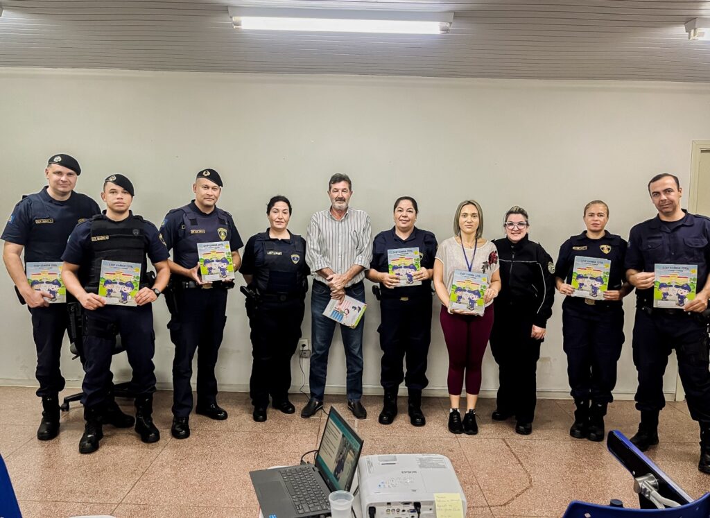 A imagem mostra um grupo de policiais militares e civis, homens e mulheres, em uniforme, segurando livros infantis. Eles estão alinhados em duas fileiras, com um homem de camisa social no meio. Há um projetor e um laptop na frente deles, sugerindo uma apresentação ou treinamento. O ambiente parece ser uma sala de aula ou de reunião. Todos parecem sorrir ou ter expressões neutras, indicando uma situação formal, mas amigável. Os livros que eles seguram têm a logo de uma organização, possivelmente a que eles pertencem, e parecem ser materiais educativos para crianças. A imagem transmite a ideia de uma iniciativa de leitura ou de conscientização infantil conduzida pela polícia.