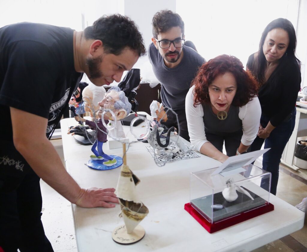 A imagem mostra um grupo de quatro pessoas, três homens e uma mulher, reunidos em torno de uma mesa branca. Eles parecem estar examinando com interesse alguns bonecos ou figuras em miniatura que estão sobre a mesa. O homem à esquerda, vestindo uma camiseta preta, está inclinado sobre a mesa, olhando para as figuras. Ele tem barba e cabelo escuro e encaracolado. Ao seu lado, um homem com óculos e barba, vestindo uma camiseta cinza, também observa as figuras. No centro, uma mulher com cabelo ruivo e encaracolado, vestindo uma blusa branca e cinza, está agachada, olhando atentamente para uma das figuras. Atrás dela, uma mulher com cabelo escuro e liso, vestindo uma blusa preta, também se inclina para ver as figuras. As figuras na mesa parecem ser caricaturas de pessoas famosas, com cabeças grandes e traços exagerados. Uma delas, com cabelo grisalho e vestida de terno, está em pé sobre uma base azul. Outra figura, que parece ser uma freira, está mais ao fundo. Há também uma figura em uma caixa de acrílico com uma base vermelha. O ambiente parece ser um escritório ou um espaço de exposição.