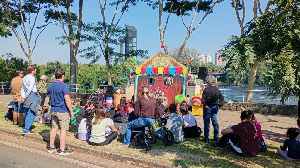 A imagem mostra uma apresentação de teatro de fantoches ao ar livre, em um parque arborizado, com um público composto por adultos e crianças. O palco é um trailer adaptado, com decoração colorida e chamativa, e a apresentação parece atrair a atenção de todos os presentes. As pessoas estão sentadas na grama, algumas em família, outras em grupos menores, e a atmosfera é descontraída e alegre. Ao fundo, observa-se um rio e prédios, indicando um ambiente urbano próximo à natureza. O dia está ensolarado e o céu está azul e limpo. Há uma mistura de pessoas de diferentes idades e, aparentemente, de diferentes grupos familiares ou sociais, unidas pela experiência compartilhada da apresentação. O ambiente é tranquilo e agradável, ideal para um evento cultural ao ar livre.