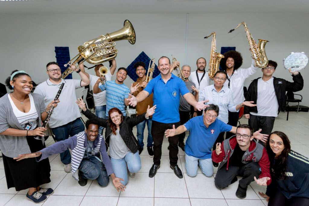 A imagem mostra um grupo grande e animado de músicos, muitos segurando seus instrumentos, sorrindo para a câmera. Há uma mistura de instrumentos de sopro, incluindo trompetes, saxofones, e um tuba. Um homem de camisa azul está no centro da foto, com os braços abertos, como se estivesse conduzindo ou liderando o grupo. As pessoas ao redor dele parecem ser de diversas idades e etnias. A atmosfera é descontraída e festiva, sugerindo uma reunião de banda ou um ensaio. O fundo é simples, uma parede branca com um pouco de decoração, indicando um ambiente interno como um salão de ensaio ou uma sala de aula. A iluminação é uniforme, sem sombras fortes, contribuindo para a clareza da imagem.