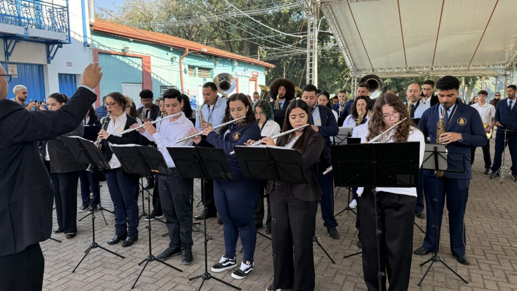 A imagem mostra uma banda de música tocando ao ar livre. A banda é composta por vários músicos, a maioria deles jovens, tocando instrumentos como flautas, clarinetes, saxofones e trompetes. Eles estão vestidos com uniformes escuros e estão posicionados em uma formação organizada. O local parece ser uma rua ou praça, com edifícios ao fundo. O clima parece ser ensolarado. Um maestro está à frente da banda, conduzindo a orquestra. Há um grande número de pessoas assistindo à apresentação, embora não sejam visíveis na imagem. A cena é vibrante e cheia de energia, mostrando a paixão e a dedicação dos músicos. A imagem é bem iluminada e nítida, permitindo uma visualização clara de todos os detalhes.