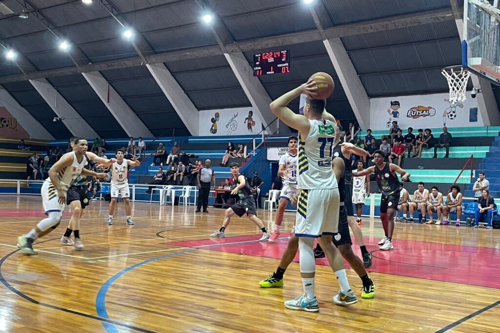 A imagem mostra uma partida de basquete em uma quadra coberta. Vários jogadores estão em ação na quadra, com um jogador em destaque no ato de arremessar a bola. Outros jogadores estão posicionados em diferentes partes da quadra, alguns defendendo e outros se preparando para receber a bola. As arquibancadas estão parcialmente ocupadas por espectadores assistindo ao jogo. O placar eletrônico mostra o resultado da partida, indicando que o jogo está em andamento. A iluminação da quadra é artificial e bem distribuída, proporcionando boa visibilidade do jogo. A quadra parece bem conservada e limpa. No fundo, há uma parede com grafites relacionados ao esporte. O ambiente geral transmite a atmosfera de uma partida de basquete amadora ou semiprofissional.