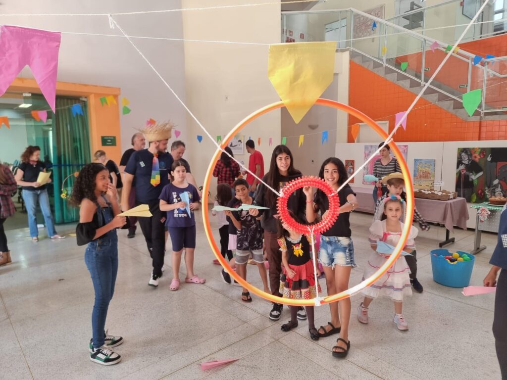A imagem mostra uma festa junina em um ambiente interno, provavelmente uma escola ou salão de festas. Crianças e adultos estão reunidos em torno de uma estrutura circular laranja, que parece ser parte de uma brincadeira ou jogo. As crianças estão segurando aviõezinhos de papel e algumas estão usando roupas típicas de festa junina. Há bandeirinhas coloridas penduradas no teto, criando uma atmosfera festiva. O ambiente é alegre e descontraído, com pessoas sorrindo e interagindo. No fundo, há uma escada e algumas paredes com quadros. A cena sugere uma celebração animada e participativa de uma festa junina.