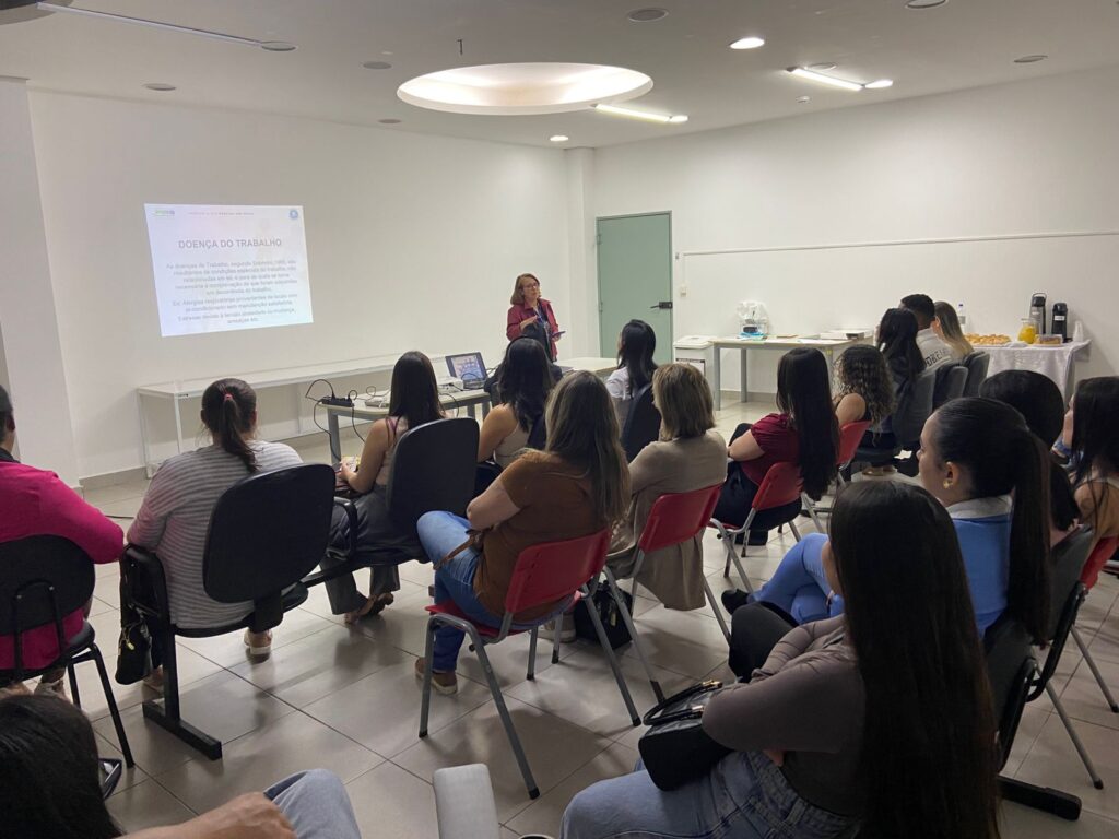 A imagem mostra uma sala de palestras com uma apresentação em andamento sobre "Doença do Trabalho". Uma palestrante está na frente, apresentando o conteúdo em um projetor. A plateia é composta por várias mulheres sentadas em cadeiras vermelhas, prestando atenção à apresentação. Há um lanche leve disponível em uma mesa lateral. O ambiente é formal e profissional, típico de uma sala de treinamento ou conferência. A iluminação é suave e o clima parece ser tranquilo e focado na apresentação. A imagem captura um momento de aprendizado e compartilhamento de informações sobre Acidentes de Trabalho e Relatório de Atendimento ao Acidentado do Trabalho - RAAT