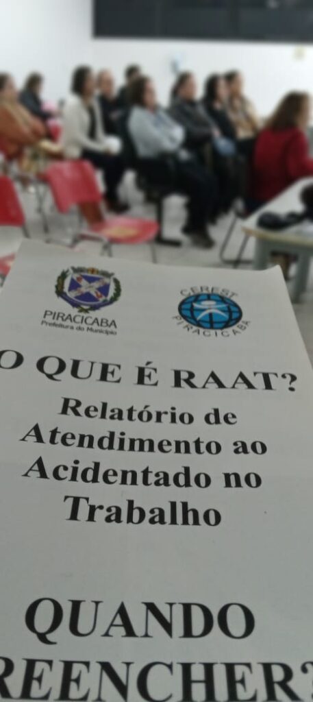 A imagem mostra um documento impresso com o título "O QUE É RAAT?". Abaixo do título, está escrito "Relatório de Atendimento ao Acidentado no Trabalho".  Há também a pergunta "QUANDO PREENCHER?" na parte inferior. No topo do documento, há os logotipos da Prefeitura Municipal de Piracicaba e do CEREST Piracicaba. Ao fundo, observa-se uma sala de aula ou auditório com pessoas sentadas em cadeiras, ligeiramente desfocadas, sugerindo que a foto foi tirada durante uma apresentação ou palestra. As cadeiras são vermelhas e estão dispostas em fileiras. 