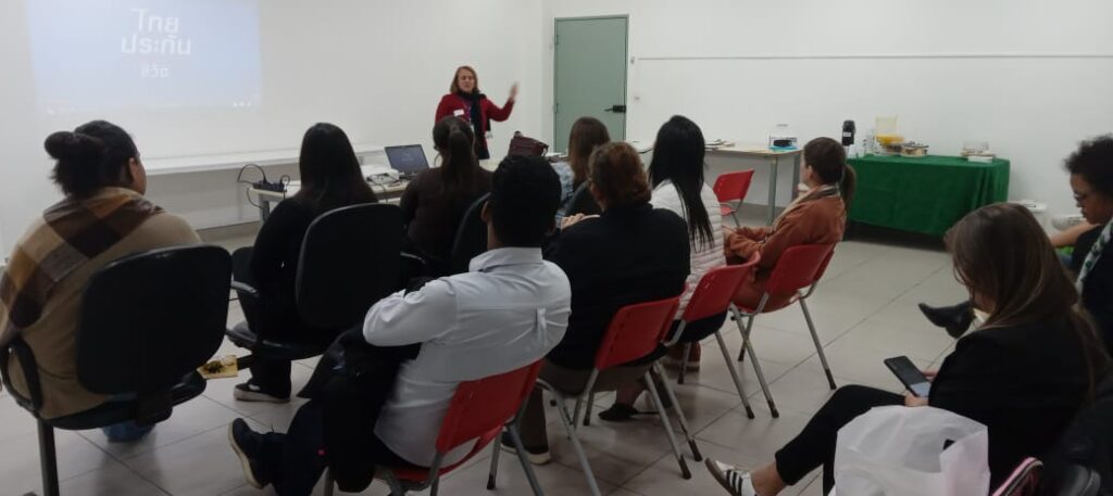 A imagem mostra uma sala do Hospital Unimed com uma apresentação em andamento. Uma palestrante do CEREST está de pé na frente de uma tela de projeção, gesticulando enquanto fala para um grupo de pessoas sentadas em cadeiras vermelhas. A sala é simples, com paredes brancas e um chão claro. Há uma mesa com alguns objetos sobre ela, possivelmente materiais para a apresentação. As pessoas na plateia parecem estar atentas à palestrante, algumas tomando notas ou interagindo entre si.