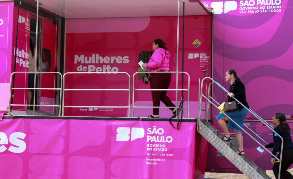 A imagem mostra um caminhão-móvel rosa vibrante, parte de uma campanha de saúde pública chamada "Mulheres de Peito" do governo do estado de São Paulo. O caminhão tem uma escada de acesso e várias mulheres estão interagindo com ele. Uma mulher de blusa rosa está perto da entrada, aparentemente esperando ou participando de alguma atividade. Outra mulher, de vestido azul, sobe a escada, enquanto uma terceira, de blusa escura, está no chão próximo ao caminhão. A imagem é bem iluminada e mostra detalhes claros do caminhão e das pessoas presentes. A repetição da frase "São Paulo, Governo do Estado, São Paulo para todos" e o logotipo do governo reforçam a mensagem da campanha.