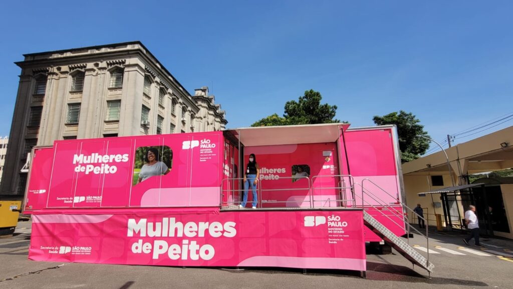 A imagem mostra um caminhão-móvel rosa vibrante, com a frase "Mulheres de Peito" escrita em letras grandes e brancas. O caminhão parece ser uma unidade móvel de saúde, possivelmente para exames de mama. Há imagens de mulheres em janelas cortadas no caminhão, e uma mulher de máscara está em pé na plataforma de acesso. O caminhão está estacionado em frente a um prédio de estilo clássico e há pessoas no fundo. O logotipo do governo de São Paulo está presente no caminhão. A imagem sugere uma campanha de saúde pública voltada para o público feminino, focando na prevenção e detecção precoce do câncer de mama.