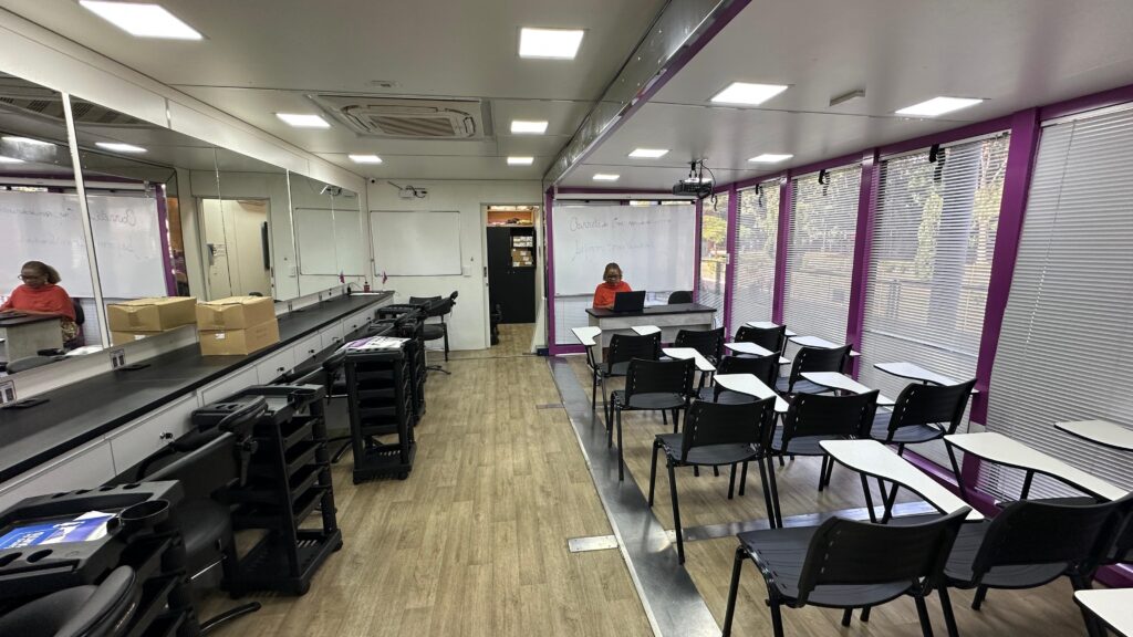 A imagem mostra uma sala de aula ou salão de beleza dentro de um contêiner. Há uma parede com espelhos ao longo de um lado da sala, com várias estações de trabalho equipadas com carrinhos de beleza. Do outro lado da sala há carteiras e cadeiras organizadas em fileiras, voltadas para uma mesa onde uma mulher está sentada em um laptop. Há uma lousa branca na parede, com algumas anotações escritas. A sala tem janelas com persianas e iluminação artificial. O chão é de madeira e o ambiente parece limpo e organizado. A atmosfera geral sugere um espaço de aprendizagem ou treinamento profissional.