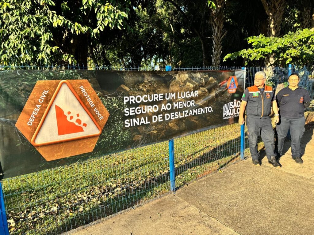 A imagem mostra uma faixa grande com um aviso de deslizamento de terra. A faixa está em português e tem um triângulo de advertência com pedras caindo. Há texto que diz "Defesa Civil", "Protege você", "Procure um lugar seguro ao menor sinal de deslizamento", e o logotipo do governo do estado de São Paulo. Duas pessoas uniformizadas de Defesa Civil estão em pé ao lado da faixa. A faixa está fixada em uma cerca em um ambiente externo com vegetação. A imagem parece ter sido tirada durante o dia com boa iluminação.