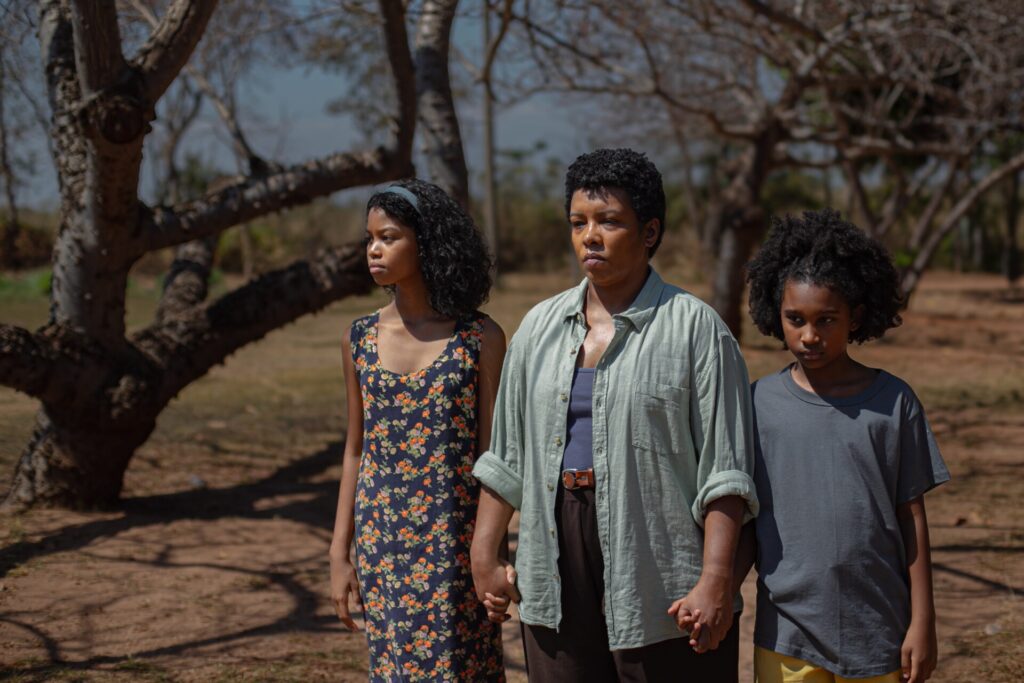 A imagem mostra três mulheres negras caminhando juntas em uma trilha de terra. A mulher do meio, que parece ser a mais velha, está usando uma camisa verde-acinzentada e calças escuras. Ela está segurando as mãos das duas mulheres mais jovens, uma de cada lado. A mulher mais jovem à esquerda está usando um vestido floral e a mulher mais jovem à direita está usando uma camiseta cinza e shorts amarelos. Todas as três mulheres parecem estar olhando para a frente com expressões sérias. O cenário é uma área arborizada com árvores nuas ou quase nuas, sugerindo que pode ser outono ou inverno. O solo é seco e poeirento. A iluminação sugere que é dia, com o sol brilhando sobre as mulheres. A atmosfera geral da imagem é calma, mas também um tanto melancólica. A composição da imagem é simples, mas eficaz. As três mulheres são o foco principal, e o fundo desfocado ajuda a destacar suas figuras. A paleta de cores é principalmente terrosa, com os tons verdes, cinzas e marrons do cenário contrastando com os tons mais escuros das roupas das mulheres. A imagem como um todo evoca uma sensação de unidade familiar e resiliência, mesmo diante de um cenário potencialmente difícil.