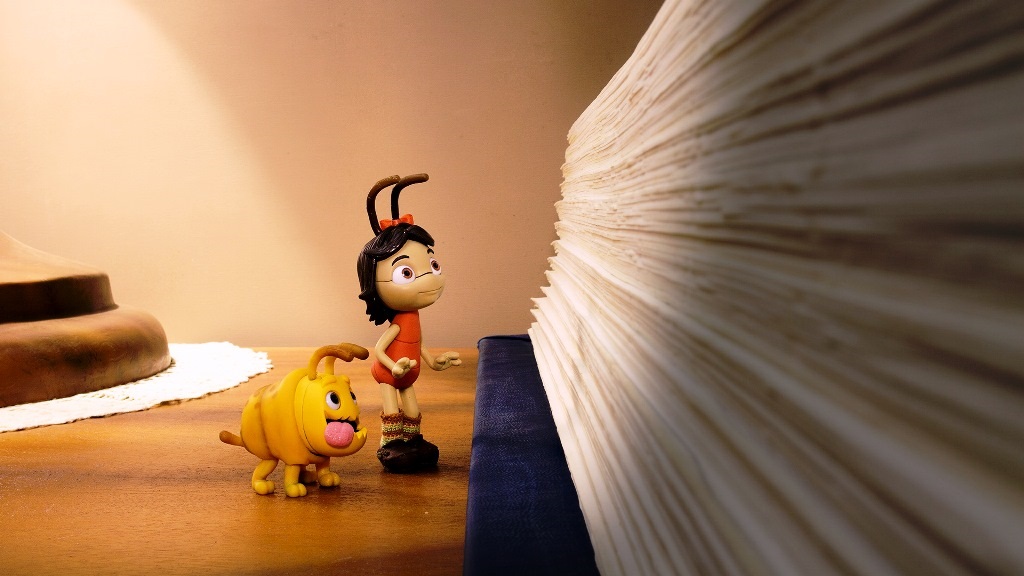 A imagem mostra duas figuras pequenas, uma formiga antropomórfica feminina e um cachorro amarelo, em uma mesa de madeira. A formiga, com um vestido laranja e meias verdes, está de pé perto de uma pilha alta de livros, olhando para ela com curiosidade. O cachorro está sentado ao lado da formiga. O fundo é uma parede bege clara, e uma lâmpada está parcialmente visível no canto superior esquerdo. A cena é bem iluminada, com a luz da lâmpada criando um brilho suave. A imagem parece ser uma foto de uma cena de brinquedo ou modelo, com uma qualidade quase surrealista. A profundidade de campo é rasa, concentrando-se nas figuras e deixando a pilha de livros levemente desfocada no fundo.