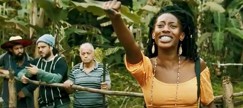 A imagem mostra um grupo de pessoas em uma floresta tropical. Uma mulher negra com dreadlocks e um vestido laranja está no centro da imagem, com o braço levantado em um gesto de protesto ou celebração. Ao seu redor, há três homens e uma mulher mais velha. Os homens parecem ser de diferentes idades e etnias, e estão vestindo roupas simples. A mulher mais velha está usando uma camisa listrada. O fundo é uma floresta tropical exuberante, com muitas árvores e plantas verdes. A cena parece ser uma captura de um filme ou programa de televisão, e a atmosfera é tensa e dramática. A imagem sugere uma luta pela terra ou pelos direitos humanos.