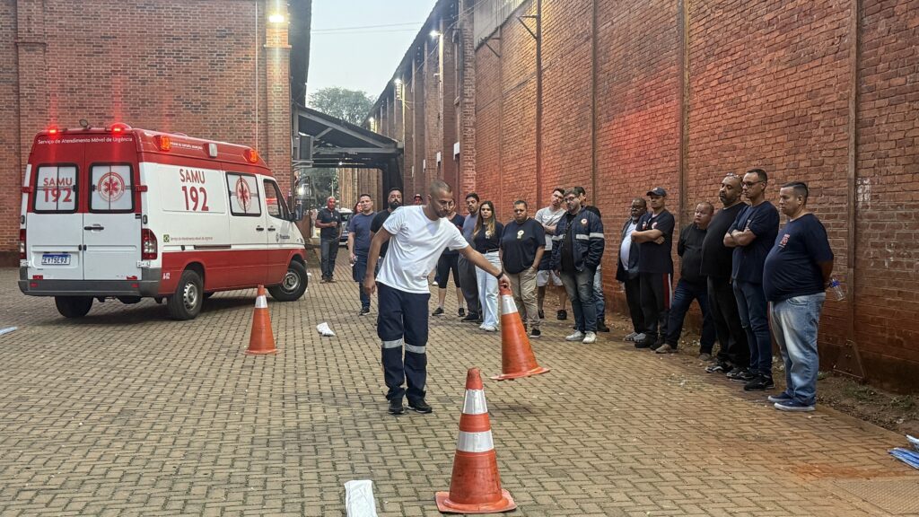 Um grupo de pessoas está reunido ao ar livre, com uma ambulância exibida de forma proeminente no lado esquerdo da imagem. A ambulância é branca e vermelha, com "SAMU 192" escrito em seu lado e na parte de trás, indicando que faz parte do Serviço de Atendimento Móvel de Urgência. As luzes de emergência do veículo estão piscando em vermelho. No primeiro plano, um homem com uma camiseta branca e calças azuis com listras refletivas está interagindo com cones de sinalização. Ele parece estar demonstrando ou montando uma área. Atrás dele, uma linha de homens está parada, alguns com os braços cruzados, observando a cena. Eles estão vestidos com roupas casuais, sendo alguns com camisetas escuras e outros em trajes mais formais. O fundo consiste em um grande edifício de tijolos com uma passagem coberta ou estrutura se estendendo a partir dele. O chão é pavimentado com ladrilhos. A impressão geral é de um encontro ao ar livre ou sessão de treinamento relacionada aos serviços de emergência.