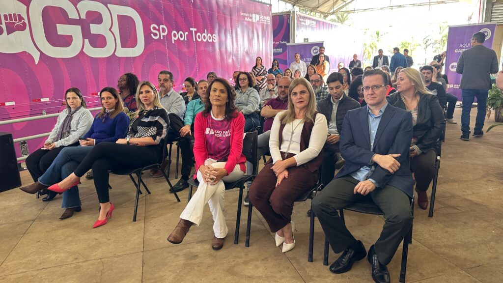 A imagem mostra uma reunião de mulheres em cadeiras, sentadas em frente a um caminhão decorado com o logotipo "GBD SP por Todas". As mulheres parecem estar participando de um evento ou conferência. Há também homens presentes no fundo, alguns sentados e outros em pé. O ambiente parece ser externo, sob uma estrutura coberta. O tom geral da imagem é formal, com as mulheres vestindo roupas elegantes. A imagem é bem iluminada e nítida. A imagem sugere um evento relacionado a mulheres e seus direitos, dado o logotipo no caminhão.