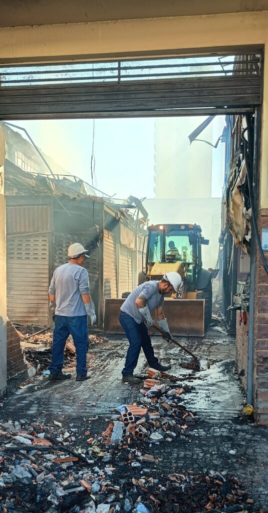 A imagem mostra uma cena de destruição após um incêndio. Duas pessoas, vestindo capacetes e luvas de proteção, estão no que parece ser uma rua ou passagem entre edifícios, limpando escombros. Uma delas usa uma pá para recolher detritos, enquanto a outra observa. Ao fundo, um trator com uma pá carregadeira está posicionado, sugerindo que a limpeza e remoção de entulho estão em andamento. Os edifícios ao redor parecem ter sofrido danos significativos pelo fogo, com estruturas parcialmente desabadas e sinais visíveis de queimadura. Fumaça ainda pode ser vista subindo em direção ao céu claro. A cena geral transmite uma sensação de devastação e o início do processo de recuperação.