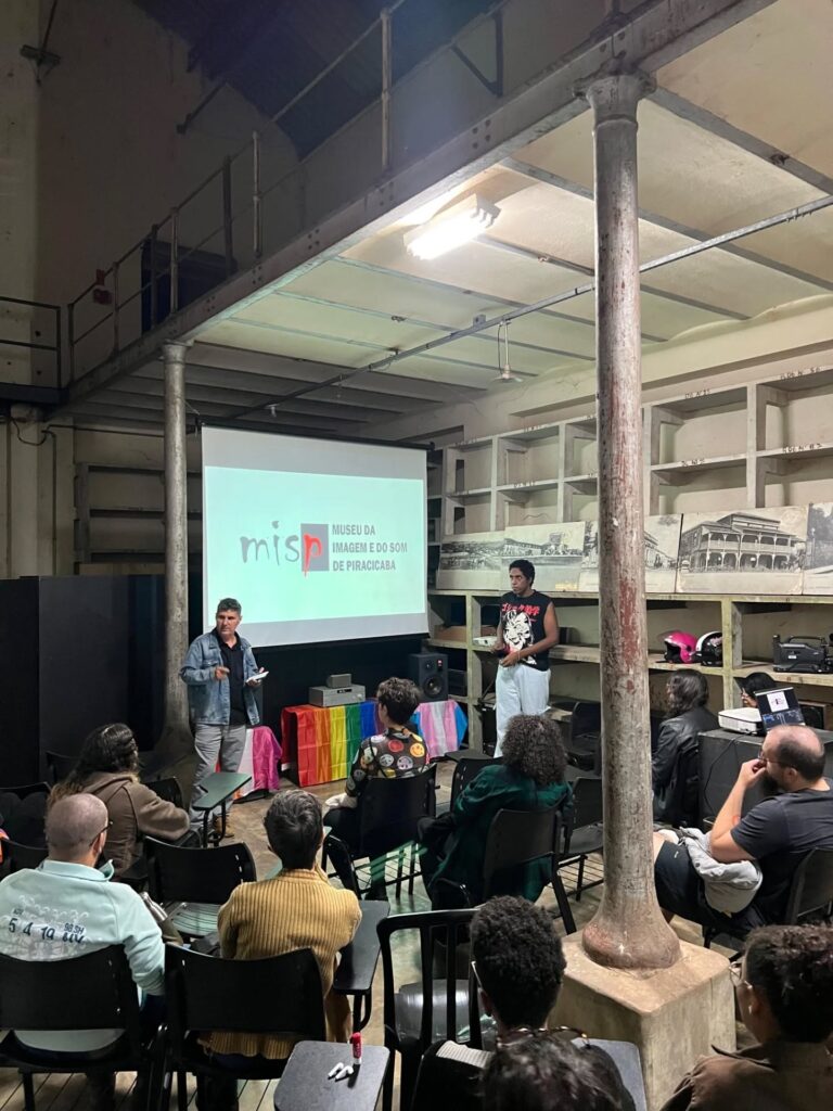 A imagem mostra uma palestra ou apresentação num espaço industrial convertido. O local parece ser um armazém ou fábrica antigo, com colunas de suporte de ferro fundido expostas, pisos de cimento e prateleiras de metal nas paredes. As prateleiras contêm vários itens, incluindo fotografias em preto e branco que parecem ser de arquivo histórico. Uma tela de projeção mostra o logotipo de um museu, “MISP Museu da Imagem e do Som de Piracicaba”. Dois apresentadores estão de pé em frente à tela, um homem e uma mulher, ambos aparentemente a falar para uma audiência sentada em cadeiras escolares pretas. A audiência é composta por várias pessoas, com roupas casuais e diversificadas. Há uma bandeira arco-íris visível no chão, sugerindo um evento LGBTQIA+ ou inclusivo. A iluminação é baixa, com a maior parte da luz vindo do projetor e de lâmpadas fluorescentes no teto. A atmosfera geral é informal, mas focada na apresentação. A imagem sugere um evento comunitário ou educacional num espaço cultural alternativo.