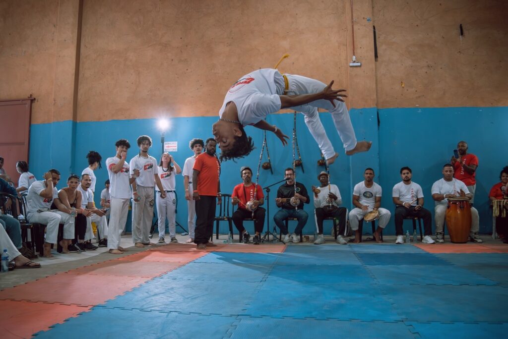 A imagem mostra uma roda de capoeira em um ginásio. No centro, um capoeirista realiza um movimento aéreo, um salto mortal, com o corpo arqueado e os braços abertos. Ele veste uma roupa branca com detalhes amarelos, que parecem ser de um cordão de capoeira. Ao redor, espectadores e músicos compõem a cena. Um grupo de pessoas está sentado em cadeiras, alguns segurando instrumentos musicais como berimbau, pandeiro e atabaque, que são típicos da capoeira. Outras pessoas estão em pé, observando a apresentação. O ginásio possui paredes com pintura desgastada, combinando tons de laranja e azul, e o chão é coberto por tatames nas cores azul e vermelho. Uma iluminação forte, possivelmente de um refletor, destaca a área central da roda.