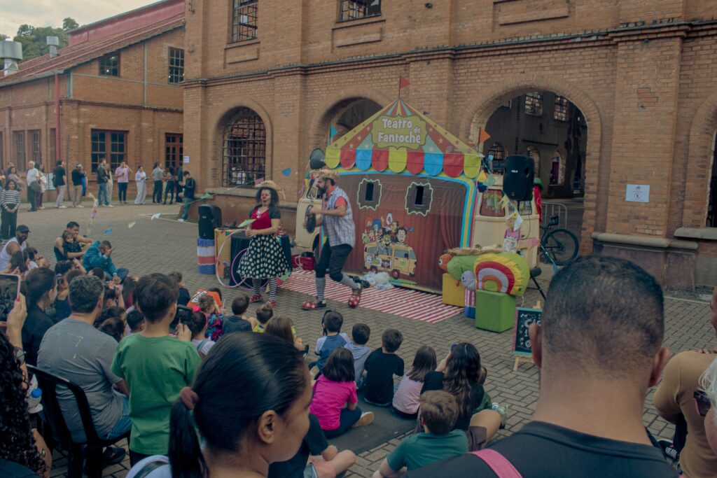 A imagem mostra uma apresentação de fantoches ao ar livre, com uma tenda colorida ao fundo e dois artistas vestidos de palhaços. Uma multidão de pessoas, incluindo muitas crianças, está sentada no chão, assistindo à apresentação. O cenário é uma área pavimentada em frente a um edifício de tijolos com arcos, sugerindo um local público ou um centro cultural. Há decorações como bandeirinhas e balões, e alguns objetos cênicos como um grande bicho de pelúcia.