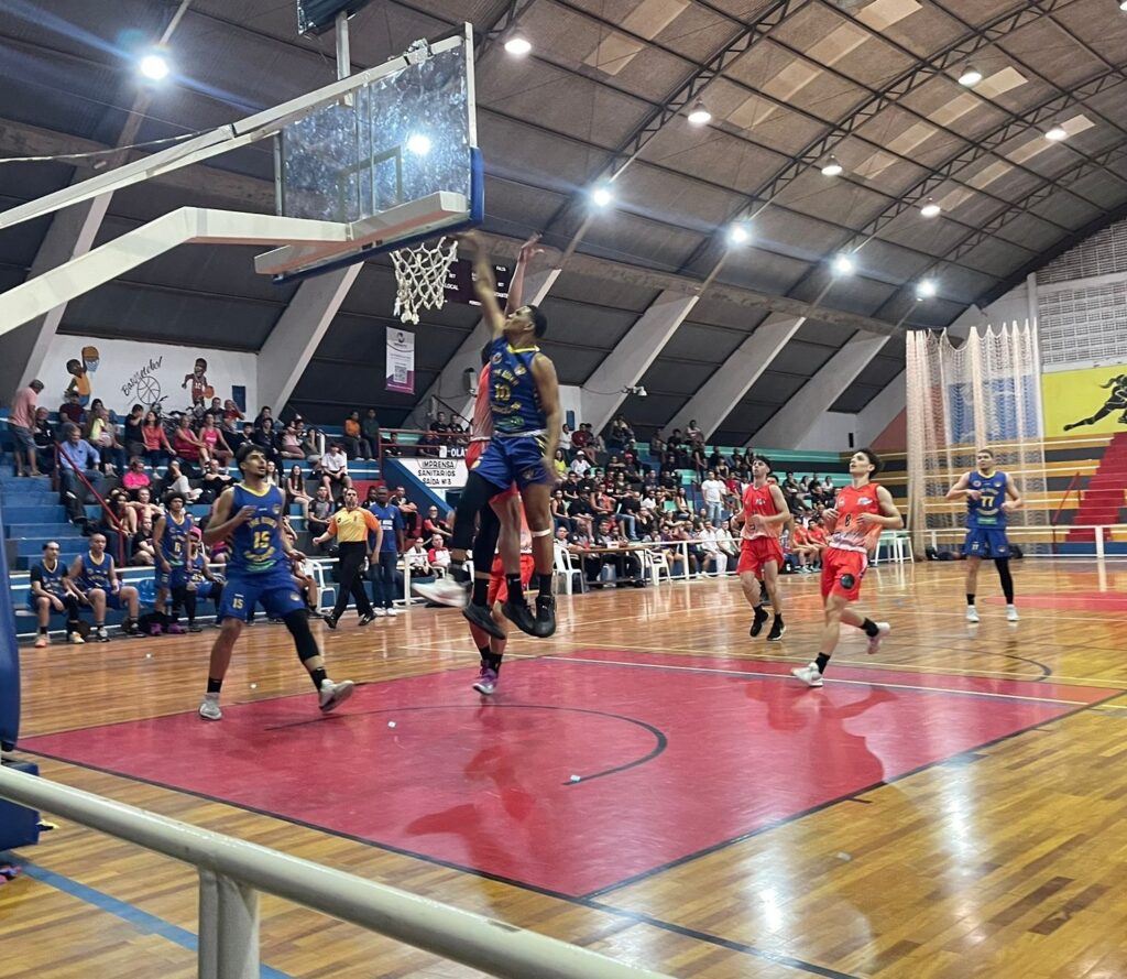 A imagem mostra uma partida de basquete em uma quadra interna. No centro da imagem, um jogador de uniforme azul e amarelo está no ar, prestes a marcar uma cesta. Ele está sendo marcado de perto por um jogador de uniforme vermelho. Outros jogadores, tanto do time azul quanto do time vermelho, estão espalhados pela quadra, alguns observando a jogada, outros se posicionando. Ao fundo, arquibancadas lotadas de espectadores assistem à partida. As arquibancadas são azuis e há pessoas sentadas em diferentes níveis. A quadra possui piso de madeira com marcações vermelhas e amarelas. A iluminação é feita por lâmpadas no teto. Há também cartazes e decorações nas paredes, incluindo um desenho de um jogador de basquete.