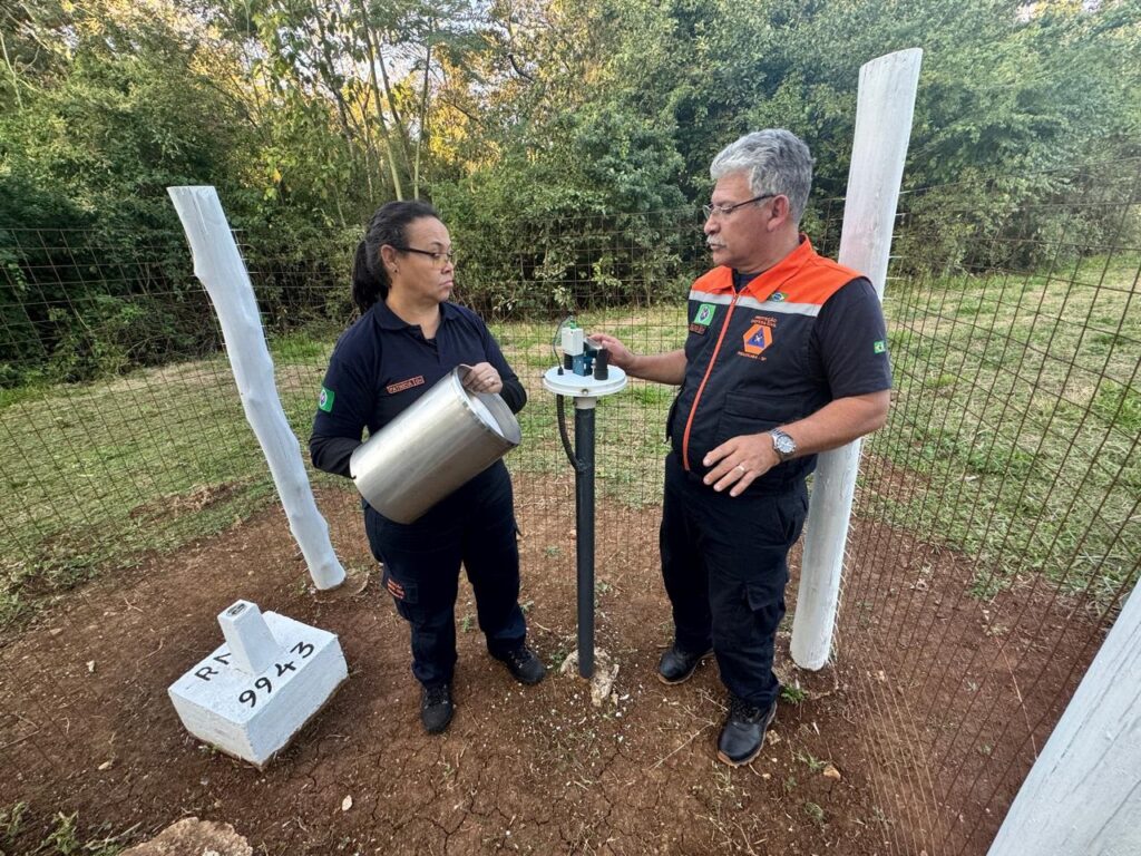 A imagem mostra dois indivíduos, um homem e uma mulher, aparentemente envolvidos em uma atividade de monitoramento ou coleta de dados meteorológicos. A mulher, vestindo uniforme escuro com um logo, segura um recipiente metálico, possivelmente para coletar amostras de água ou outro material. O homem, também uniformizado, com um colete laranja e preto com logotipos, parece estar operando ou inspecionando um equipamento instalado em um poste metálico. O equipamento tem sensores e parece ser um instrumento meteorológico. Ao redor, há uma cerca de arame e postes de madeira brancos. No chão, há uma base de concreto com inscrições, possivelmente indicando uma localização ou código. O ambiente parece ser externo, em uma área com vegetação. A imagem sugere uma atividade profissional relacionada à meteorologia ou monitoramento ambiental.