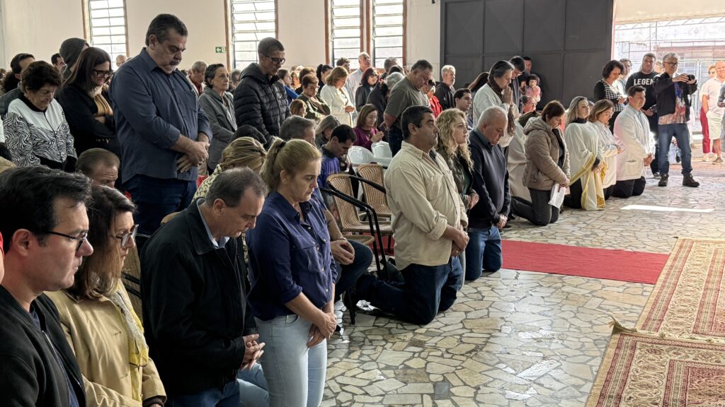 A imagem mostra um grande grupo de pessoas ajoelhadas em oração dentro de um salão amplo e iluminado. As pessoas estão vestidas com roupas casuais, e a maioria parece estar em silêncio e concentrada. Há um tapete vermelho no chão na frente do grupo, e um tapete ornamental mais elaborado perto da câmera. O salão tem paredes claras e janelas grandes que permitem que a luz natural entre. A atmosfera é calma e respeitosa, sugestiva de um evento religioso ou memorial. O fundo mostra algumas pessoas em pé, observando a cena. Há uma mistura de idades e gêneros entre os presentes. A imagem é nítida e bem iluminada, permitindo a visualização de detalhes faciais e expressões das pessoas.