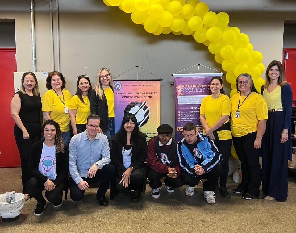 A imagem mostra um grupo de pessoas em pé e sentadas em frente a um banner. O banner é amarelo e preto e tem informações sobre o Centro de Doenças Infectocontagiosas (CEDIC) e testes rápidos para hepatites. As pessoas na imagem estão vestindo camisetas amarelas. Algumas estão sentadas no chão, outras em pé atrás delas. Há balões amarelos atrás do grupo. A imagem parece ter sido tirada em um evento ou conferência.