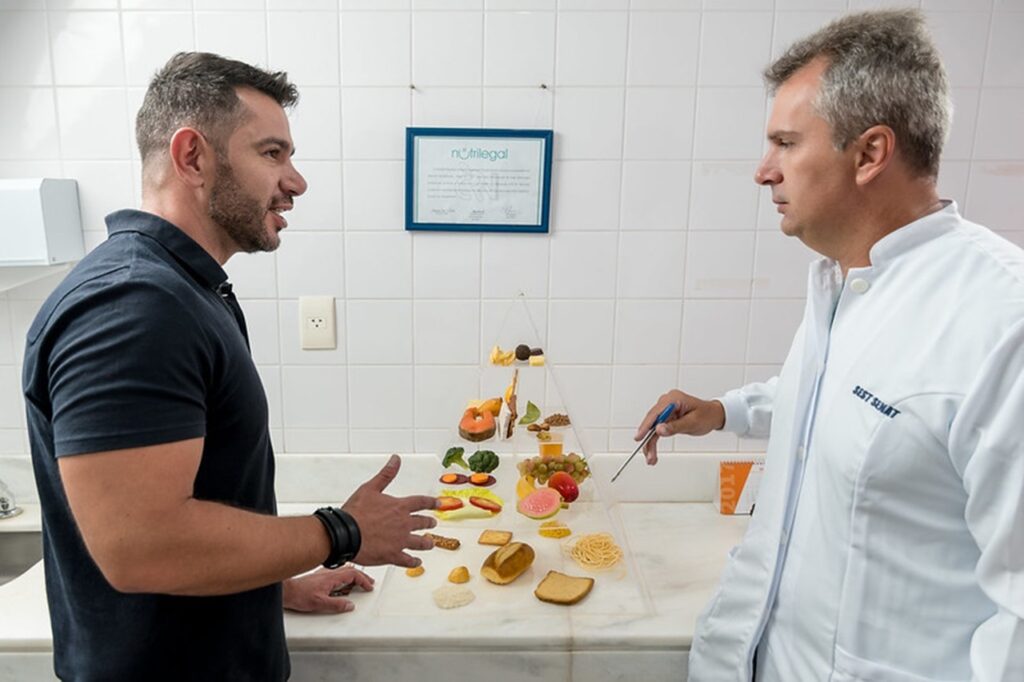 A imagem mostra dois homens em uma conversa, aparentemente numa consulta médica ou nutricional. Um deles está vestindo uma camisa polo preta e o outro um jaleco branco com o nome "Scott Schat" bordado. Entre eles, há uma pirâmide de alimentos construída com modelos de comidas, representando um guia alimentar. Ao fundo, há um certificado emoldurado na parede. A cena se passa em um ambiente limpo e claro, com azulejos brancos. A expressão corporal dos homens sugere uma discussão focada e engajada sobre o guia alimentar.