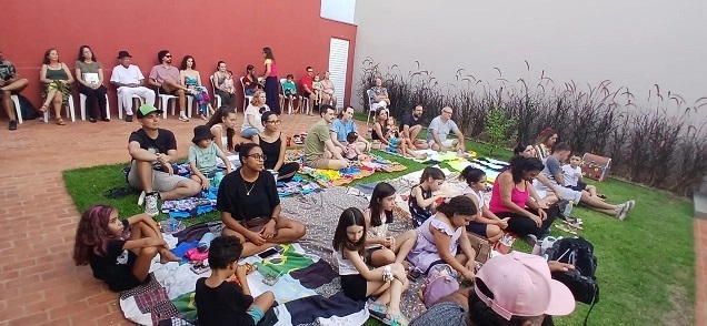 A imagem mostra um grupo de pessoas reunidas ao ar livre, sentadas em toalhas no gramado, em frente a uma parede vermelha e um jardim com grama alta. Parece ser um evento social ou uma apresentação, pois há pessoas sentadas em cadeiras ao fundo e outras no chão, prestando atenção em algo à frente. As pessoas no gramado estão sentadas em diferentes arranjos, algumas em grupos menores, outras mais espalhadas. Há uma mistura de adultos e crianças. As toalhas no chão são coloridas e parecem ser usadas para acomodar as pessoas confortavelmente. Ao fundo, algumas pessoas estão sentadas em cadeiras brancas, observando o evento. A arquitetura ao redor sugere que o local pode ser um pátio ou um espaço comunitário. A iluminação indica que é durante o dia, possivelmente no final da tarde. O ambiente geral é relaxado e informal.
