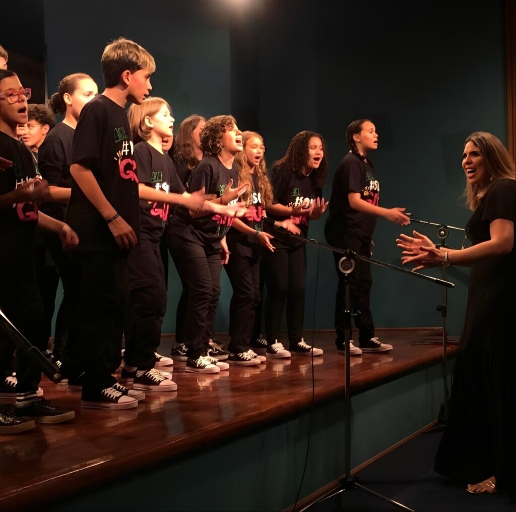 A imagem mostra um grupo de crianças e adolescentes se apresentando em um palco, possivelmente um coral ou um grupo de teatro musical. Eles estão vestidos com camisetas pretas com uma estampa colorida e calças pretas, e todos usam tênis brancos com detalhes pretos. As crianças estão posicionadas em degraus, com alguns microfones à frente delas. Elas parecem estar cantando com entusiasmo, com expressões faciais animadas e gestos com as mãos. Uma mulher adulta, vestida de preto, está em pé à direita da imagem, de frente para as crianças, e parece estar regendo ou dirigindo a apresentação, com as mãos levantadas em um gesto de encorajamento ou instrução. O palco tem um piso de madeira brilhante, e o fundo é uma parede azul escura. A iluminação parece ser focada no grupo, criando um ambiente de performance. A atmosfera geral é de energia e alegria, sugerindo uma apresentação bem-sucedida.